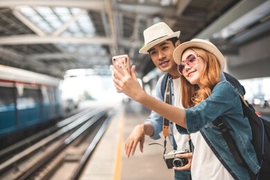 happy asian couple traveler holding a mobile phone in station and waiting for train in vacation time.two asian tourists with backpacks train travel in sightseeing city thailand.