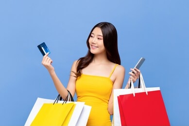 young smiling beautiful asian woman making online payment through mobile phone via credit card while carrying colorful shopping bags in isolated blue background