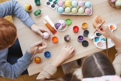 high angle view of two friends sitting together at the table and painting easter eggs with paintbrushes