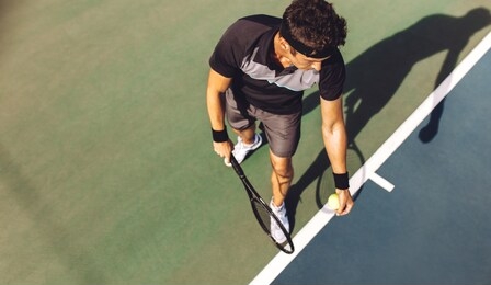 top view of a young tennis player with racket ready to serve a tennis ball. professional player standing at the baseline holding the tennis racket and the ball on a hard court.