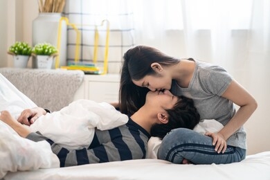 asian pretty woman sitting on bed bending face down and kissing boyfriend lying on her leg, looking on eyes and smile feeling love very much. romantic moment of couple lover in valentine day concept.