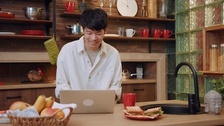 smiling young man sit and use laptop work in the morning at home in kitchen portrait male morning asia asian casual chinese guy, social media internet notebook pc close up slow motion