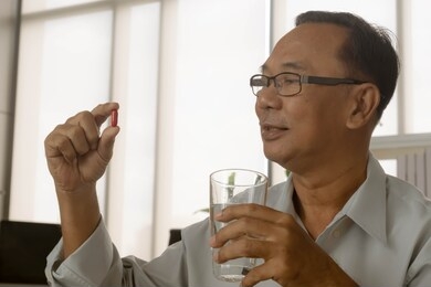 senior asian man taking red pills with water glass at home.