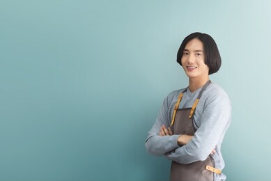 happy smiling asian man barista in brown apron with arms crossed looking to camera and standing over blue or green background. copy space. business owner.