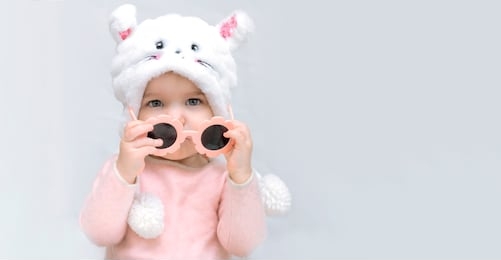 little girl in fluffy hat holds sunglasses in pink dress on white gray background. pastel natural colors. fashion concept for kids, children s style, change of seasons. banner copy space