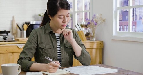 young elegant house wife writing on notebook and calculating on calculator about debt bill monthly at dining table in home kitchen. korean woman managing expense payroll. money risk financial concept
