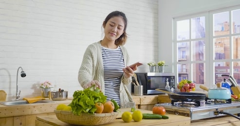 young happy asian japanese smiling woman wear sweater looking recipe in mobile phone while standing in kitchen. vegetable salad dieting healthy lifestyle concept. girl cooking at home prepare food