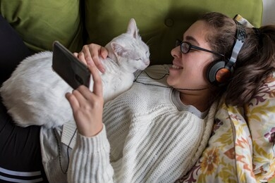 young girl lying on a sofa watching a video on the mobile, with white cat on her chest. young girl with glasses and headphones, white sweater.