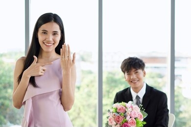a young beautiful asian woman with happy face showing a wedding ring, dating with a man with smiling face in black suit holding a pink rose bouquet in the restaurant