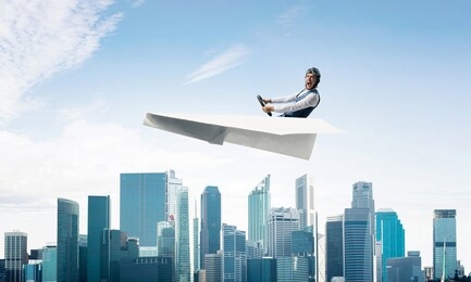 scared pilot with open mouth sitting in paper plane and holding steering wheel. aviator driving paper plane above business center in blue sky. cityscape with high skyscrapers and office buildings.