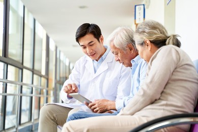 young asian doctor discussing test result and diagnosis with senior couple patients using digital tablet in hospital hallway
