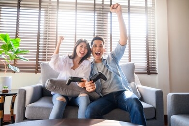 happy asian couple using joystick controller playing video games together while sitting on sofa  in their living room