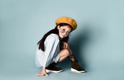 little asian child in sunglasses, shirt dress, brown beret, boots. she smiling, posing cross-legged sitting on floor against blue background. childhood, fashion, hipster style. close up, copy space
