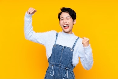 young asian girl in overalls over isolated yellow background celebrating a victory
