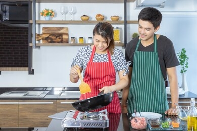 happy beautiful asian wife is cooking for breakfast while husband stands beside to help and prepare food in modern kitchen in holidays. couple is smiling, feeling in love. valentines day concept.