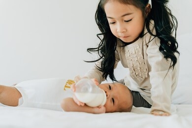 asian family of cute little sister feeding bottle of milk to newborn baby boy brother. toddler kid and new sibling relax in a white bedroom at home