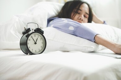 black vintage retro alarm clock times at 1 o'clock and tablet on bed with happy young asian pretty woman in casual clothing sleeping at home in background, healthy sleep and joyful weekend concept