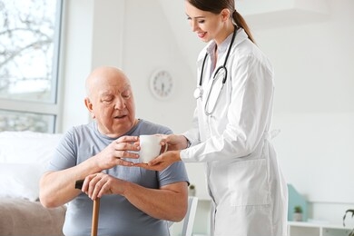 female doctor giving senior man suffering from parkinson syndrome cup of tea in clinic