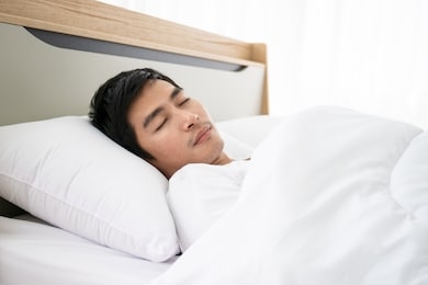 handsome asian young man talking pleasure in sleeping on the bed in morning. around twenties years old man resting on the bed in bedroom. asian ethnicity man lying down on the bed and wake up late.