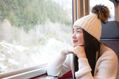 an asian woman is sitting happily looking at the natural view from the window on the train and traveling on a vacation in switzerland and planning a train trip.