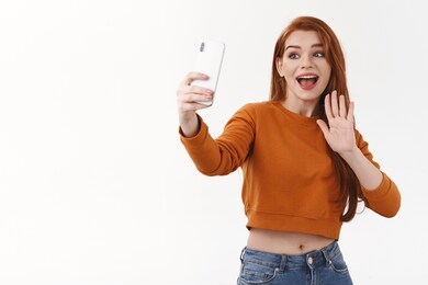 friendly excited happy redhead redhead woman in orange cropped sweater, talking with granny via smartphone online video-call, connect to friends chat, waving palm say hello or hi, talking followers