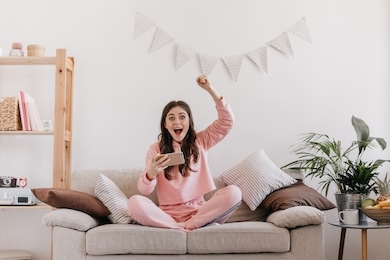 joyful girl in sports outfit is sitting on sofa, holding phone and emotionally enjoying victory. surprised woman in pink shirt playing games on smartphone at home