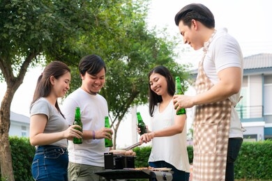 asian man cooking barbeque grill and sausage for a group of friends to eat party in garden at home. group of friends having outdoor garden barbecue laughing with alcoholic beer drinks