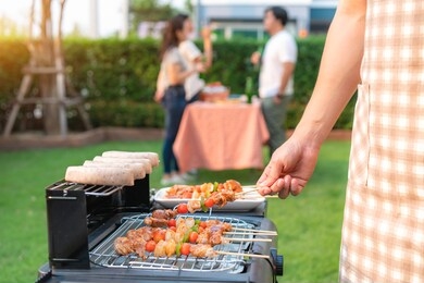 asian man cooking barbeque grill and sausage for a group of friends to eat party in garden at home. group of friends having outdoor garden barbecue laughing with alcoholic beer drinks