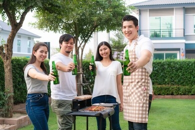 asian man cooking barbeque grill and sausage for a group of friends to eat party in garden at home. group of friends having outdoor garden barbecue laughing with alcoholic beer drinks
