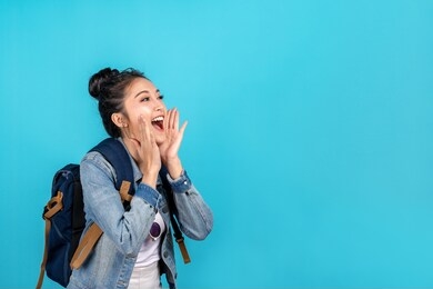happy asian woman travel backpacker shouting open mouth to copyspace on blue background. cute asia girl smiling wearing casual jeans shirt and looking to aside for present promotions.