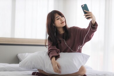 asian happy woman sitting on bed is enjoying listening to music and calling video from her smartphone with earphones. technology connection digital online marketing ecommerce banner concept.