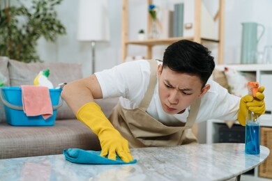 husband housekeeping and cleaning concept. concentrated young asian chinese man in yellow rubber gloves wiping dust using spray and duster on table in home living room. frowning guy doing housework