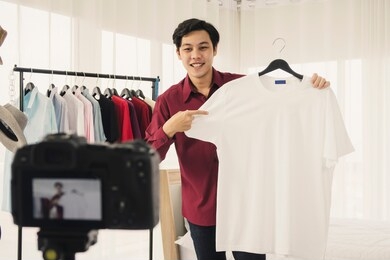 happy young attractive asian male blogger or vlogger present and review t-shirt on live streaming broadcast with internet subscribers. social media influence people, content maker, e-commerce concept.