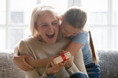 happy school girl granddaughter cuddling hugging embracing from back excited surprised middle aged retired grandmother with gift box in hands head shot. mothers day, birthday celebration concept.