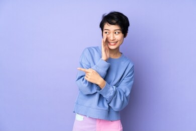 young vietnamese woman with short hair over isolated purple background pointing to the side to present a product and whispering something