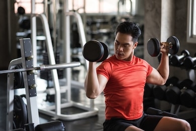 strong asian muscle man exercise biceps with two dumbbells with gym equipment background. muscular body sportsman workout in sport fitness center. bodybuilding and healthy lifestyle concept.