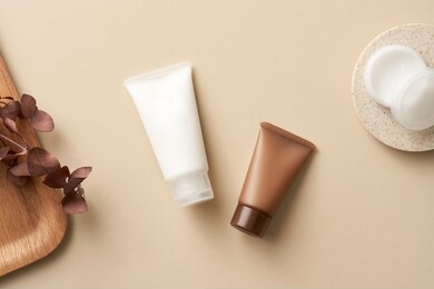 top view composition of white and brown plastic tubes for cosmetic products placed on beige surface with open bottle with white cream and wooden tray with plant