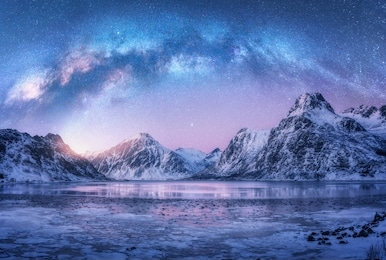 milky way above frozen sea coast and snow covered mountains in winter at night in lofoten islands, norway. arctic landscape with blue starry sky,  water, ice, snowy rocks, milky way. space and galaxy