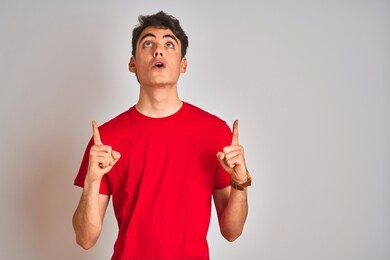 teenager boy wearing red t-shirt over white isolated background amazed and surprised looking up and pointing with fingers and raised arms.