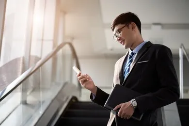 a asian handsome young businessman holding his laptop tablet while using his phone and standing on the escalator. business concept.