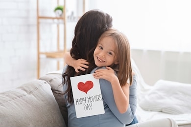 smiling little daughter with happy mother's day gift card hugging her mom, celebrating international mother's day together at home