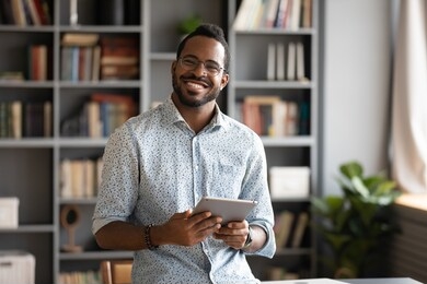 smiling young biracial man in glasses holding digital computer tablet, lookig at camera. addicted to technology, happy handsome businessman using electronic device, web surfing internet indoors.
