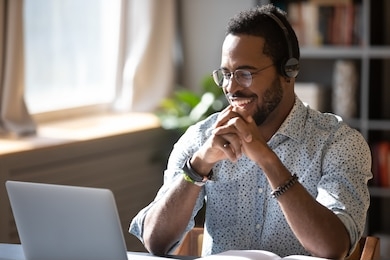 happy millennial african american man in glasses wearing headphones, enjoying watching educational webinar on laptop. smiling young mixed race businessman holding video call with clients partners.