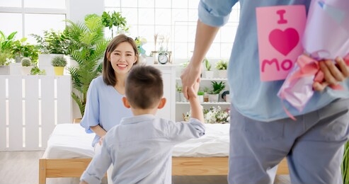 happy family - father and son are hiding flowers and card for their mother behind backs while she is sitting at home