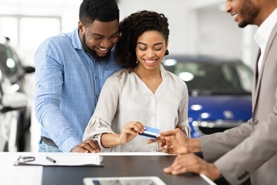 buying car concept. smiling black spouses giving credit card to automobile seller in auto selling showroom. selective focus