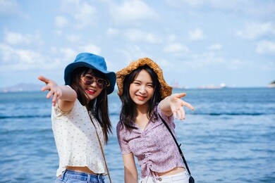 two girls enjoy tpgether at the beach while they travel at the sea, reach their hands to the camera.