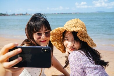 two girls sitting on the beach at the sea in sunny day, take a selfie together and making duck face.