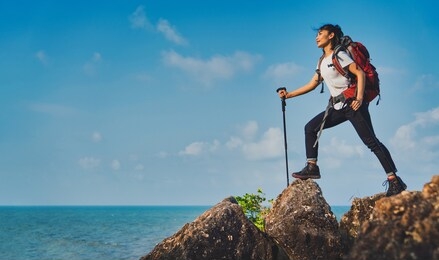young asian women hikers climbing up on the peak of mountain near ocean. woman hiking in the mountains standing on a rocky summit ridge with backpack and pole looking out over ocean landscape