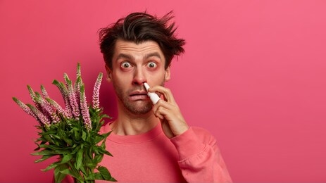 sick young man has allergic reaction to flowering plant, uses nasal spray to cure rhinitis, has red swelling eyes, symptoms of allergy, unhealthy look, sneezes all time. spring and healthy problems