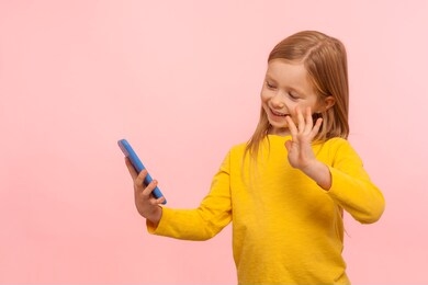 portrait of happy beautiful little girl with ginger hair smiling and gesturing hi hello to mobile phone, child communicating with parents on video call. indoor studio shot isolated on pink background